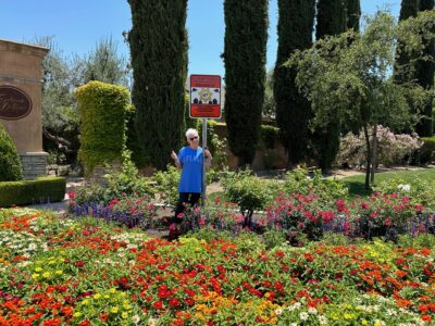Sue Kendall stands next to a neighborhood watch sign in a flower garden.